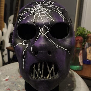 Puede incluir: Una máscara morada y negra con un diseño agrietado. La máscara tiene grandes agujeros para los ojos, una nariz y una boca con barras de metal. La máscara es probablemente para disfraces o cosplay.