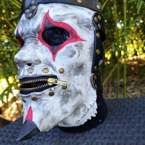Könnte beinhalten: Eine gealterte weiße Maske mit roten Akzenten um Augen und Nase. Die Maske hat einen schwarzen Reißverschluss für den Mund und wird mit einem schwarzen Lederriemen und Messingnieten befestigt. Die Maske ist zum Tragen bestimmt.