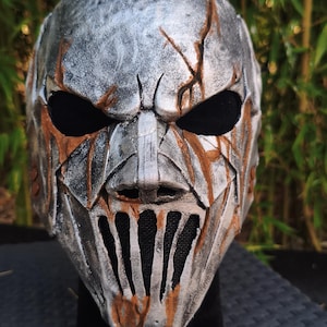 May include: A full-face mask featuring a weathered silver finish with deep brown, rust-like accents in the crevices. The mask has large, dark, hollow eye sockets and a vertical, barred mouth opening covered with black mesh fabric.