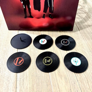Könnte beinhalten: Set aus sechs schwarzen Vinyl-Schallplatten-Untersetzern mit verschiedenen farbigen Logos. Ein Untersetzer ist ein durchgehender schwarzer Kreis mit einem Zeigerdesign. Die Untersetzer sind auf einer hellen Holzoberfläche mit rotem Hintergrund angeordnet.