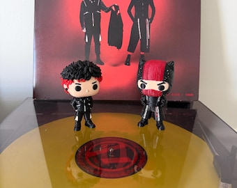 Op de afbeelding: Twee vinyl verzamelfiguren staand op een doorzichtige gele vinylplaat. Het linkerfiguur heeft zwart haar met rode accenten en een zwart jasje. Het rechterfiguur draagt een zwart-rood masker en een zwarte outfit met rode details.