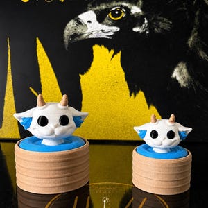 May include: Two white, horned creature figurines with large black eyes and blue side accents, each mounted on a blue circular base atop a tan, cylindrical, ribbed stand. The background features a black and yellow album cover with the text TRENCH.