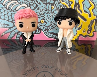 May include: Two stylized vinyl figures standing on a clear surface. The left figure has pink hair and a black coat. The right figure wears a black bucket hat, a white open coat, and gray pants. The background features a blue dragon illustration.