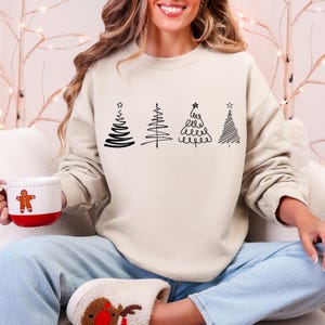 Könnte beinhalten: Ein cremefarbenes Sweatshirt mit vier schwarzen, mit Linien gezeichneten Weihnachtsbäumen. Jeder Baum hat ein einzigartiges Design, mit Sternen auf einigen. Eine rot-weiße Tasse mit einem Lebkuchenmann wird in der Hand gehalten.