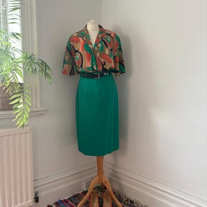 May include: A short-sleeved button-down shirt featuring an abstract pattern in shades of green, orange, and black, paired with a solid emerald green knee-length skirt and a black leather belt with a silver buckle, displayed on a mannequin.