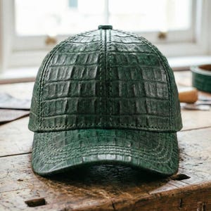 May include: A dark green baseball cap with a textured, crocodile-like pattern. The cap has a curved brim and a structured crown. The hat is made of a leather-like material and sits on a wooden surface.