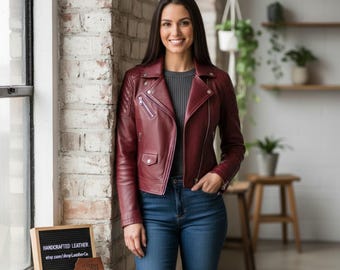 Pode incluir: Jaqueta de couro bordô com estilo motociclista clássico, com gola entalhada, fecho de zíper assimétrico e vários bolsos. A jaqueta é combinada com jeans azuis. Uma placa diz "HANDCRAFTED LEATHER" com um endereço de site.