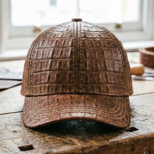 May include: A brown leather baseball cap with a textured, crocodile-like pattern. The cap features a curved brim and a stitched seam running down the center. The hat is made of a rich, brown leather.