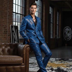 Könnte beinhalten: Ein blauer Anzug mit Schlangenmuster, bestehend aus einem Blazer und einer passenden Hose. Der Anzug wird über einem schwarzen Hemd getragen. Der Hintergrund zeigt eine Backsteinmauer, Fenster und ein Ledersofa.