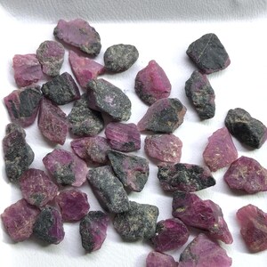 Puede incluir: Una colección de rubíes en bruto y sin pulir esparcidos sobre una superficie blanca. Cada piedra presenta una mezcla de secciones cristalinas de color rojo rosado vibrante e inclusiones minerales opacas de color gris oscuro o negro.