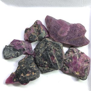 May include: A collection of six raw, unpolished ruby gemstones featuring a mix of deep magenta and dark grey, textured mineral inclusions. Each stone displays irregular, jagged edges and a natural, matte surface with varying concentrations of red.