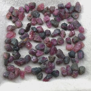 May include: A collection of numerous small, raw, unpolished gemstone fragments in varying shades of deep pink, magenta, and dark grey. These irregular, jagged mineral pieces are scattered across a textured, off-white surface, displaying natural facets.