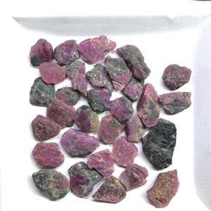 Puede incluir: Una colección de piedras preciosas de rubí en bruto y sin pulir esparcidas sobre una superficie blanca. Las piedras exhiben una mezcla de tonos magenta intenso y rosa, contrastados con inclusiones minerales oscuras, rugosas y grisáceas.