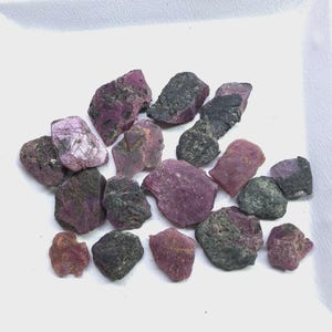 Puede incluir: Una colección de fragmentos de piedras preciosas en bruto y sin pulir en varios tonos de rosa intenso, magenta y gris oscuro. Estas piezas minerales irregulares y dentadas muestran una mezcla de superficies cristalinas y opacas.