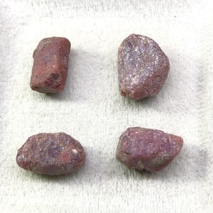 以下が含まれることがあります： 不規則で自然な形状と質感を持つ、未研磨のルビー原石4つ。それぞれの石は、深みのある土のような赤色をしており、色調や表面の粗さに微妙な変化が見られ、カットされていない鉱物標本特有の風合いを呈しています。