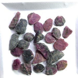 Peut inclure: Une collection de rubis bruts et non polis de tailles et de formes variées. Les pierres présentent un mélange de magenta profond, de rose et d'inclusions minérales gris foncé ou noires, révélant des textures naturelles et des bords irréguliers.