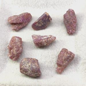 Peut inclure: Un ensemble de six pierres précieuses rubis brutes et non polies, présentant des formes irrégulières et dentelées ainsi que des surfaces texturées. Les pierres affichent des teintes rouge-rose et bordeaux, avec des formations cristallines naturelles.