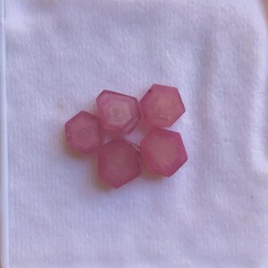 以下が含まれることがあります： 白い箱の中に、6つのピンク色の六角形の宝石が展示されています。宝石は半透明で、中央はより明るいピンク色をしています。石はクラスター状に配置され、そのユニークな形と色を示しています。