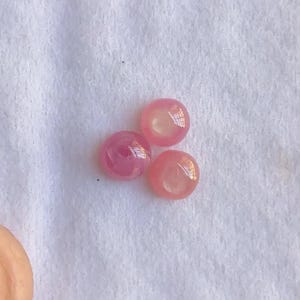 May include: Three pink, round gemstones with a glossy finish are arranged on a white, textured surface. The gems vary in shades of pink, with some appearing more translucent than others. The overall composition suggests a focus on the gems' color and texture.