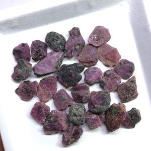 Könnte beinhalten: Eine Ansammlung roher, ungeschliffener Rubinedelsteine in verschiedenen Schattierungen von tiefem Pink und Magenta. Jeder Stein weist eine raue, unregelmäßige Textur mit dunklen, erdigen Einschlüssen und natürlichen Kristalloberflächen auf.