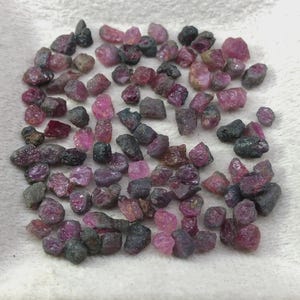 May include: A collection of small, raw, unpolished gemstones in various shades of deep pink, magenta, and dark charcoal gray. These irregular, naturally shaped mineral fragments are scattered across a textured, off-white surface.