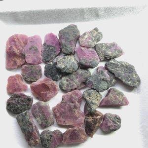 Könnte beinhalten: Eine Sammlung roher, unpolierter Rubinedelsteine mit einer Mischung aus leuchtend pink-roten kristallinen Abschnitten und einer dunklen, strukturierten grauen Mineralmatrix. Diese unregelmäßigen Mineralproben variieren.