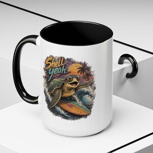 May include: A white ceramic mug with a black interior and handle, featuring a graphic of a sea turtle wearing sunglasses while surfing on a wave. The design includes a sunset, palm trees, and the text Shell yeah in a stylized orange and teal font.