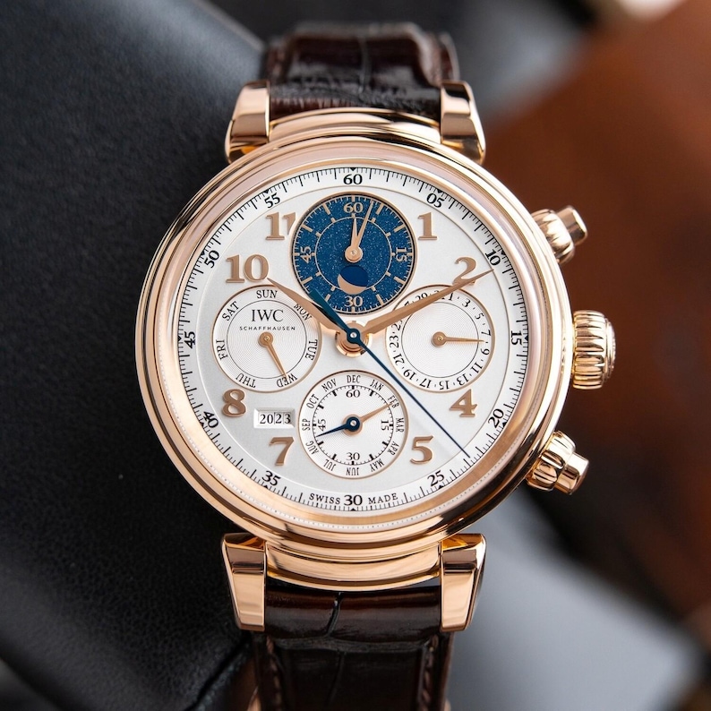 K&ouml;nnte beinhalten: Eine goldfarbene Armbanduhr mit wei&szlig;em Zifferblatt, mit mehreren Hilfszifferbl&auml;ttern und einem blauen Akzent. Die Uhr hat ein braunes Lederarmband und ist mit der Marke "IWC Schaffhausen" versehen. Die Uhr zeigt Datum und Tag an.