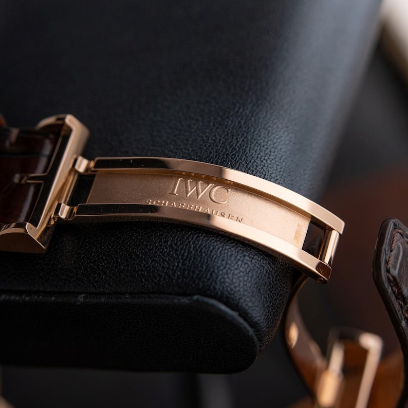 K&ouml;nnte beinhalten: Nahaufnahme einer ros&eacute;goldenen Uhrenschlie&szlig;e mit der Gravur "IWC SCHAFFHAUSEN". Die Schlie&szlig;e ist an einem braunen Lederarmband befestigt, vor schwarzem Hintergrund.