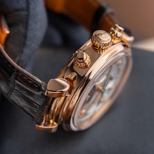 K&ouml;nnte beinhalten: Nahaufnahme einer luxuri&ouml;sen goldenen Armbanduhr mit schwarzem Lederarmband. Die Uhr hat ein detailliertes Zifferblatt und aufwendige goldene Akzente auf Geh&auml;use und Krone. Das Uhrenarmband ist dunkelbraun.