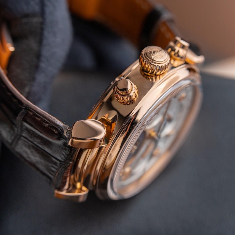 K&ouml;nnte beinhalten: Nahaufnahme einer luxuri&ouml;sen goldenen Armbanduhr mit schwarzem Lederarmband. Die Uhr hat ein detailliertes Zifferblatt und aufwendige goldene Akzente auf Geh&auml;use und Krone. Das Uhrenarmband ist dunkelbraun.