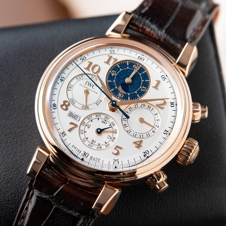 K&ouml;nnte beinhalten: Eine luxuri&ouml;se Armbanduhr mit wei&szlig;em Zifferblatt, goldfarbenem Geh&auml;use und braunem Lederarmband. Die Uhr verf&uuml;gt &uuml;ber mehrere Hilfszifferbl&auml;tter, darunter eine Mondphasenanzeige und das Datum. Die Uhr ist mit "IWC Schaffhausen" und "Swiss Made" gekennzeichnet.