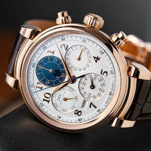 K&ouml;nnte beinhalten: Eine luxuri&ouml;se Armbanduhr mit wei&szlig;em Zifferblatt, goldfarbenem Geh&auml;use und schwarzem Lederarmband. Die Uhr verf&uuml;gt &uuml;ber mehrere Hilfszifferbl&auml;tter, darunter eine Mondphasenanzeige, sowie goldfarbene Zeiger und Markierungen. Das Zifferblatt zeigt das IWC Schaffhausen Logo.
