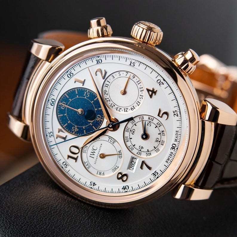 K&ouml;nnte beinhalten: Eine luxuri&ouml;se Armbanduhr mit wei&szlig;em Zifferblatt, goldfarbenem Geh&auml;use und schwarzem Lederarmband. Die Uhr verf&uuml;gt &uuml;ber mehrere Hilfszifferbl&auml;tter, darunter eine Mondphasenanzeige, sowie goldfarbene Zeiger und Markierungen. Das Zifferblatt zeigt das IWC Schaffhausen Logo.