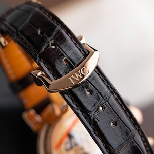 K&ouml;nnte beinhalten: Nahaufnahme eines luxuri&ouml;sen Uhrenarmbands. Das dunkelbraune Lederarmband weist ein Krokodilmuster auf und wird mit einer goldfarbenen Schnalle befestigt. Die Schnalle ist mit den Buchstaben "IWC" graviert.