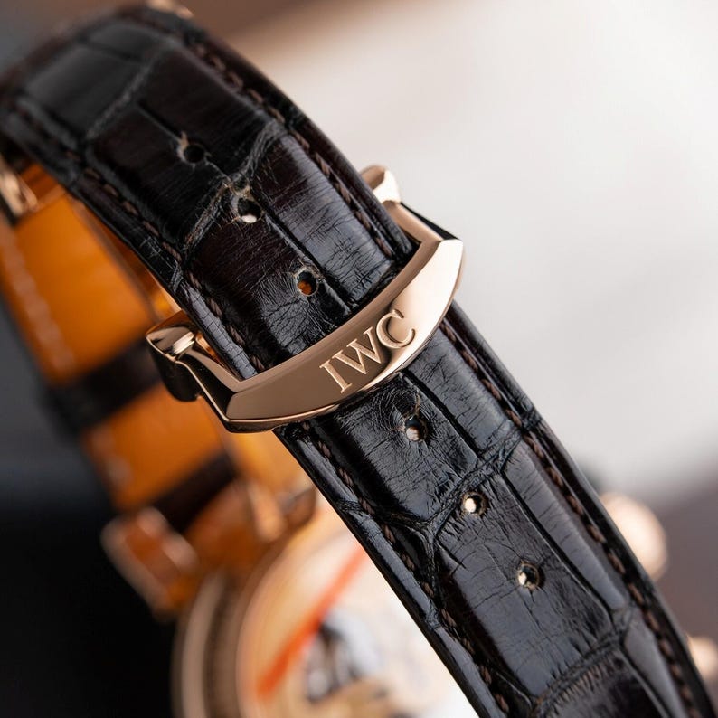 K&ouml;nnte beinhalten: Nahaufnahme eines luxuri&ouml;sen Uhrenarmbands. Das dunkelbraune Lederarmband weist ein Krokodilmuster auf und wird mit einer goldfarbenen Schnalle befestigt. Die Schnalle ist mit den Buchstaben "IWC" graviert.