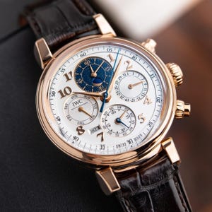 K&ouml;nnte beinhalten: Eine Luxus-Armbanduhr mit wei&szlig;em Zifferblatt, goldfarbenem Geh&auml;use und einem dunkelbraunen Lederarmband. Die Uhr verf&uuml;gt &uuml;ber mehrere Hilfszifferbl&auml;tter, darunter eine Mondphasenanzeige, und die Marke "IWC" ist sichtbar. Die Uhr hat eine Datumsanzeige und ist auf das Jahr 2023 eingestellt.