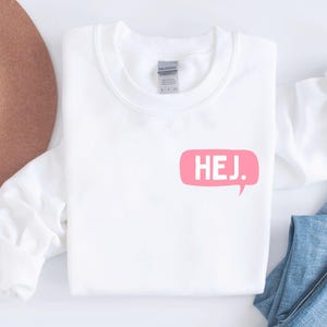 May include: A white long-sleeve crewneck sweatshirt featuring a pink speech bubble graphic on the chest with the text HEJ. printed in white inside the bubble. The sweatshirt is displayed alongside a brown wide-brim hat and blue denim jeans.