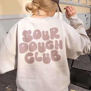 Könnte beinhalten: Ein cremefarbenes Sweatshirt mit dem Aufdruck "SOUR DOUGH CLUB" in einer Blasen-Schriftart in einem gedämpften Mauve-Ton. Das Sweatshirt wird von einer Person mit blonden Haaren und einer braunen Baseballkappe getragen.