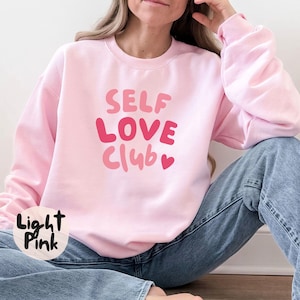 Könnte beinhalten: Hellrosa Sweatshirt mit dem Text "SELF LOVE Club" in Rosatönen. Das Sweatshirt hat einen Rundhalsausschnitt und lange Ärmel. Die Worte "Light Pink" stehen auf einem kleinen Kreis unten links.