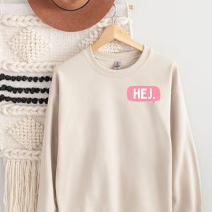 May include: A beige crewneck sweatshirt featuring a pink speech bubble graphic on the upper left chest with the white text HEJ. printed inside. The sweatshirt has long sleeves, a ribbed neckline, and a standard fit, displayed on a wooden hanger.