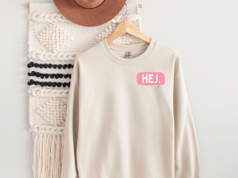 May include: A beige crewneck sweatshirt featuring a pink speech bubble graphic on the upper left chest with the white text HEJ. printed inside. The sweatshirt has long sleeves, a ribbed neckline, and a standard fit, displayed on a wooden hanger.