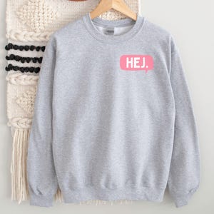 May include: A light gray crewneck sweatshirt featuring a pink speech bubble graphic on the upper left chest. Inside the speech bubble, the word HEJ. is printed in white capital letters. The sweatshirt has long sleeves and a ribbed collar.