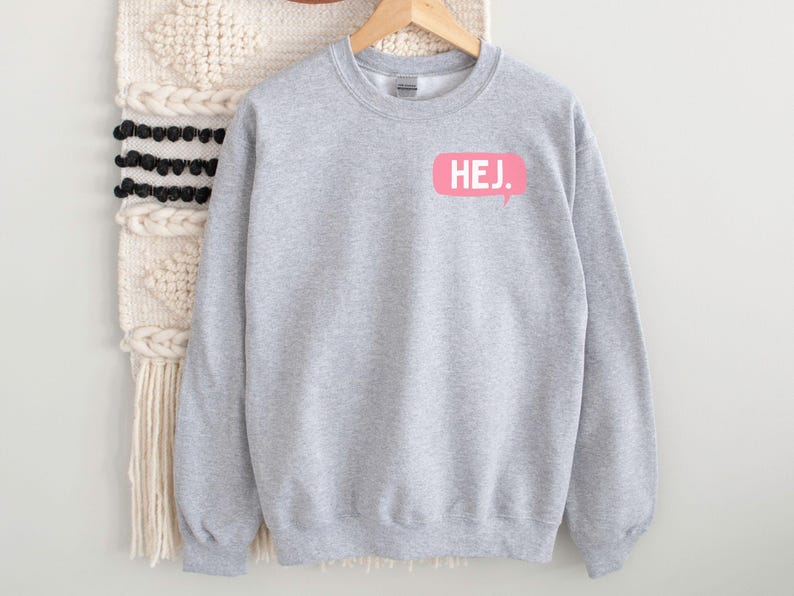 May include: A light gray crewneck sweatshirt featuring a pink speech bubble graphic on the upper left chest. Inside the speech bubble, the word HEJ. is printed in white capital letters. The sweatshirt has long sleeves and a ribbed collar.