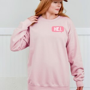 May include: A light pink long-sleeve crewneck sweatshirt featuring a small, rectangular speech bubble graphic on the upper left chest. The graphic is a darker shade of pink and contains the white text HEJ. in a clean, sans-serif font.