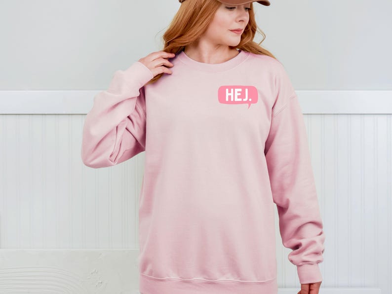 May include: A light pink long-sleeve crewneck sweatshirt featuring a small, rectangular speech bubble graphic on the upper left chest. The graphic is a darker shade of pink and contains the white text HEJ. in a clean, sans-serif font.