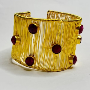 Pode incluir: Uma pulseira de punho dourada com um design de fio tecido. A pulseira apresenta várias pedras preciosas redondas, vermelho escuro. O design aberto facilita o uso e mostra o intrincado trabalho em metal. Circunferência de aproximadamente 17 cm.