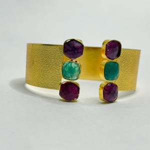 May include: Gold-tone cuff bracelet with a textured surface. The bracelet features three square gemstones in shades of purple and green on each side. The gemstones are set in gold-tone bezels.