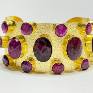 May include: A gold cuff bracelet with a textured finish, set with numerous oval and round, deep red gemstones. The bracelet's design incorporates a pattern of bezel-set stones, offering a striking and opulent appearance. The gemstones are a dark, rich red colour.