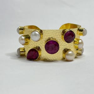 Könnte beinhalten: Goldfarbenes Armreif-Armband mit strukturierter Oberfläche, verziert mit abwechselnd runden und tropfenförmigen Rubin-Edelsteinen und weißen Perlen. Das Armband hat ein gehämmertes Metalldesign.